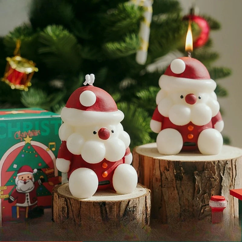 Two Santa Claus-shaped candles on wooden stumps with a Christmas tree in the background.